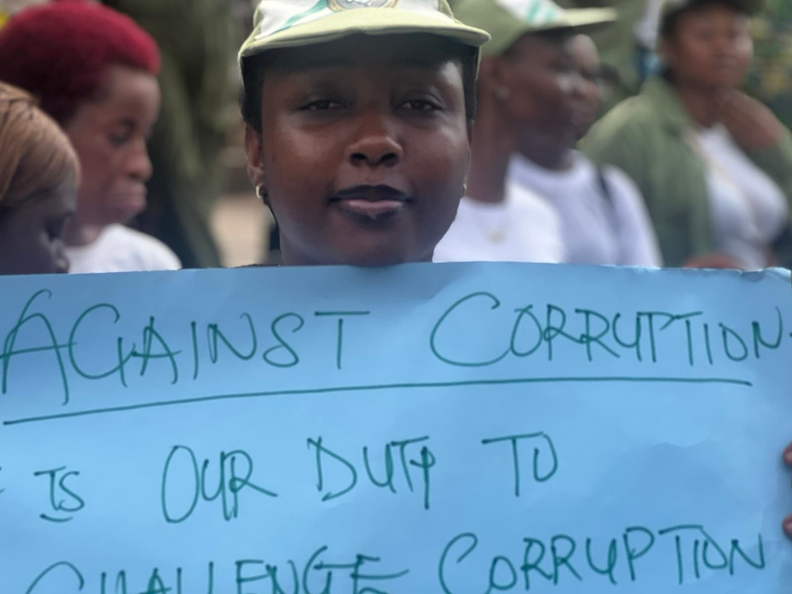National Youth Service Corps members at a community outreach, one holding a hand-lettered sign reading: against corruption — it is our duty to challenge corruption.